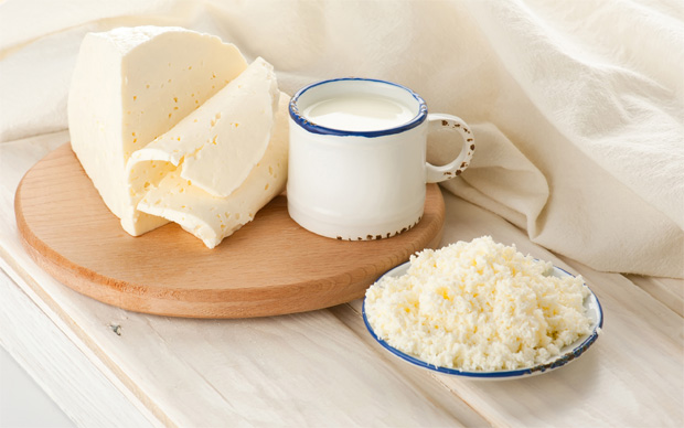 На столе круглая разделочная доска с куском сыра, кружка с молоком и миска с творогом