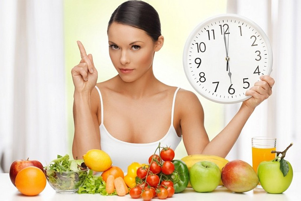На фоне полезных фруктов сидит девушка и держит большие настенные часы в руке