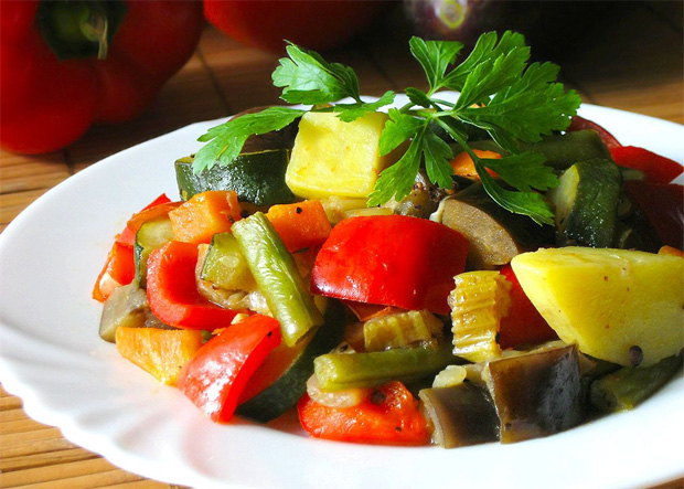 На столе в белой тарелке выложены тушеные овощи с зеленью и приправами