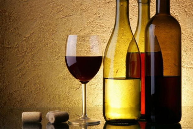 На фоне бежевой стены стоит три бутылки с вином и бокал, наполненный наполовину