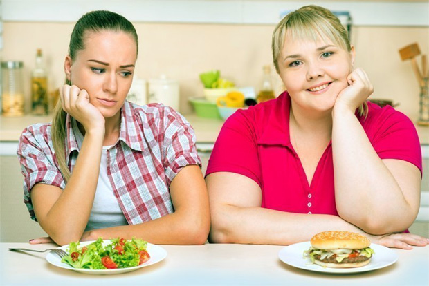 Две девушки сидят за столом с едой и худая смотрит на гамбургер у полной