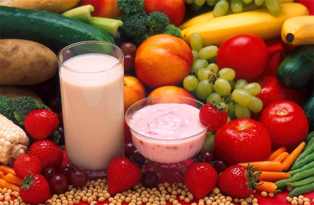На столе большое разнообразие фруктов и овощей, а также стакан с молоком и миска с йогуртом