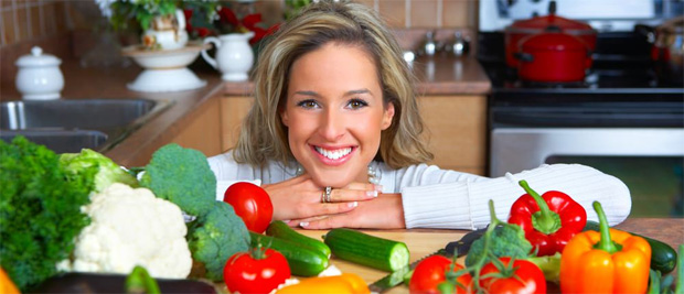 Улыбающаяся девушка стоит на своей кухне рядом с большим количеством свежих овощей
