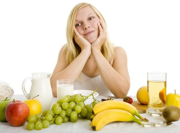 Девушка сидит за столом, оперев голову руками и задумчиво смотрит на фрукты, овощи и соки