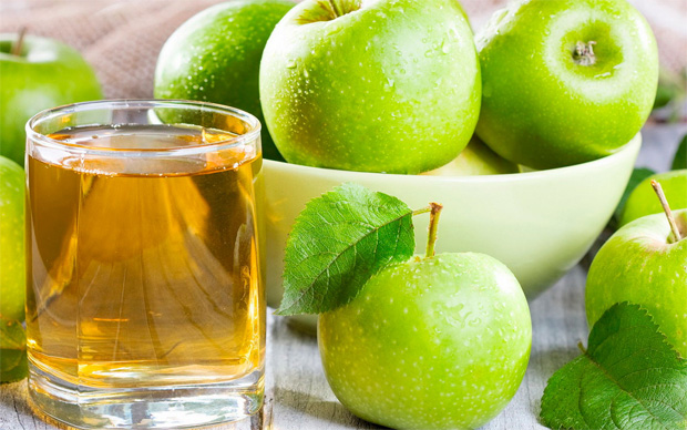 На фоне белой миски с зелеными яблоками стоит стакан с яблочным соком