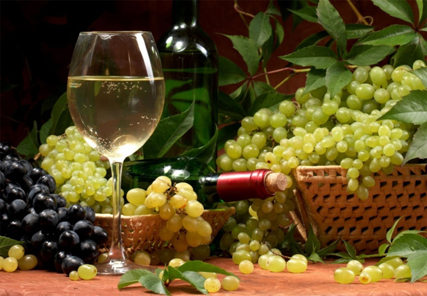 На столе большая корзина с виноградом, две бутылки и бокал с белым вином