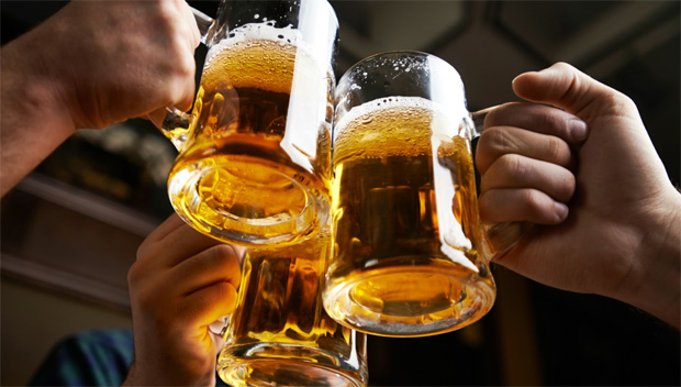 Трое мужчин в баре чокаются большими кружками с пенным пивом