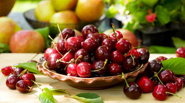 На столе на фоне ягод и фруктов стоит деревянная мисочка с ягодами черешни