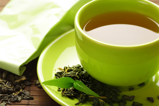 На столе в зеленой чашке с блюдцем заваренный зеленый чай и рассыпаны листья