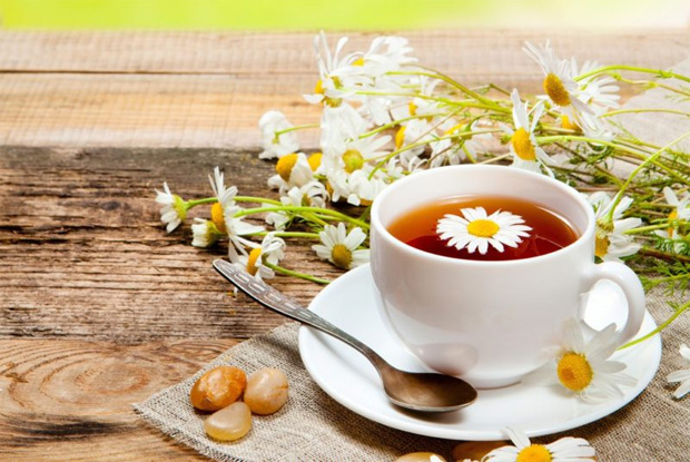 На деревянном столе стоит белая чашка с блюдцем и заваренным ромашковым чаем