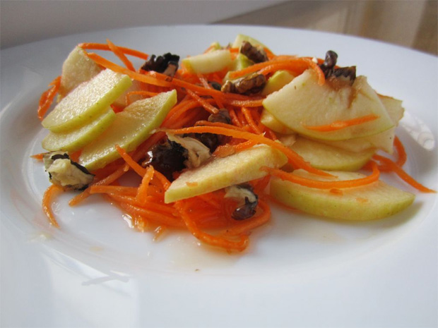 На белом блюде небольшая порция салата из яблок, моркови и орехов