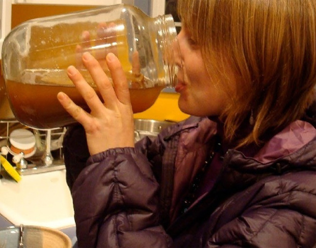 Девушка в фиолетовой куртке пьет сок чайного гриба из трехлитровой банки