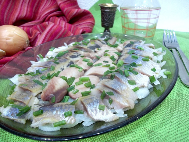 На столе большое овальное блюдо с нарезанной соленой сельдью и зеленью