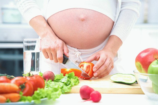 Беременная девушка на большом сроке стоит на кухне и нарезает овощи на салат