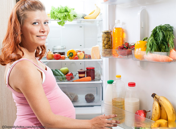 Беременная девушка стоит возле открытого холодильника на кухне и выбирает продукты