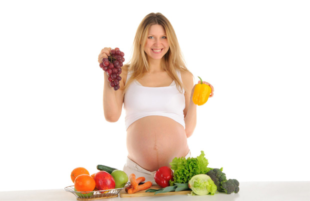В светлом помещении стоит беременная девушка у стола с фруктами и овощами