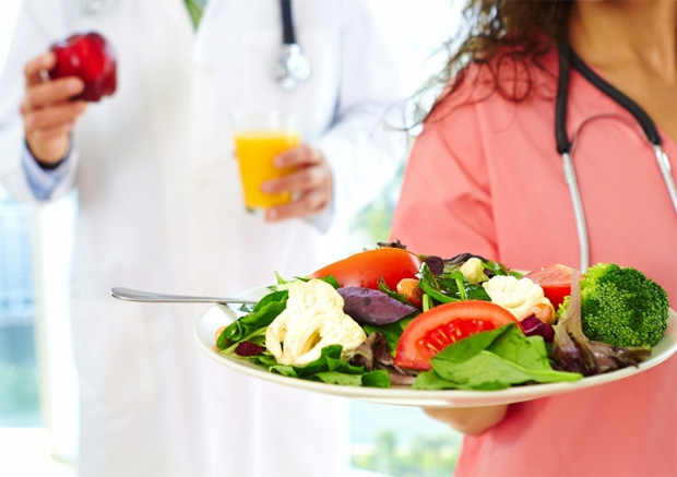 На аднем фоне врач в белом халате держит в руках овощи, а медсестра протягивает блюдо с салатом