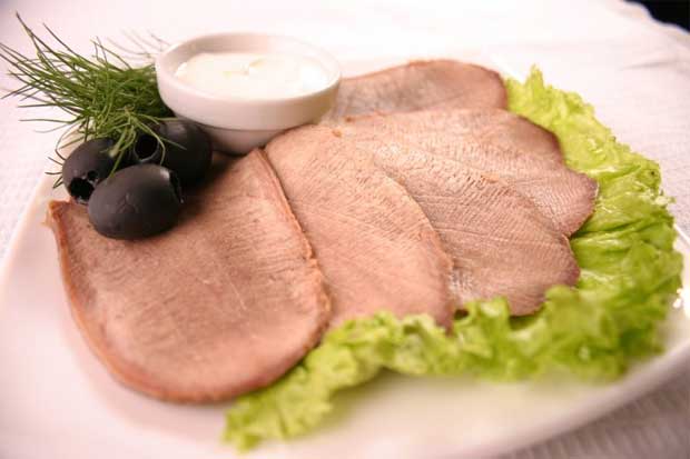На белой тарелке на листе салата выложено нарезанное мясное изделие с маслинами