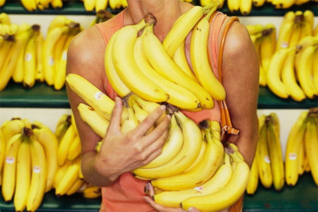 В овощном магазине женщина держит в руках несколько гроздьев бананов