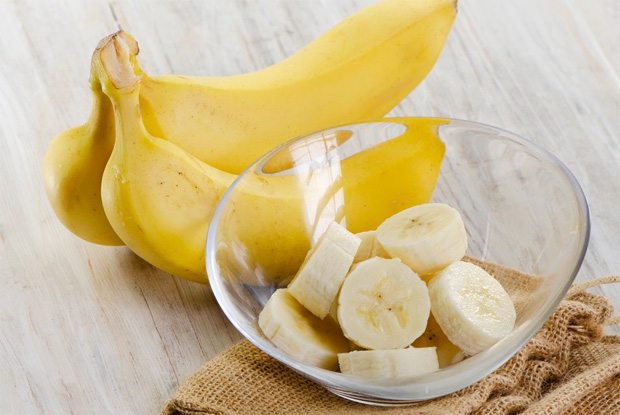 На столе лежат два целых банана и мисочка с нарезанным фруктом