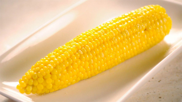 На белом блюде лежит большой початок очищенной кукурузы