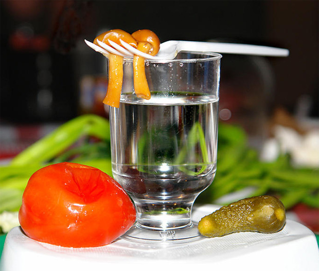 На столе стоит рюмка водки, соленые огурчик и помидор, и вилочка с грибами