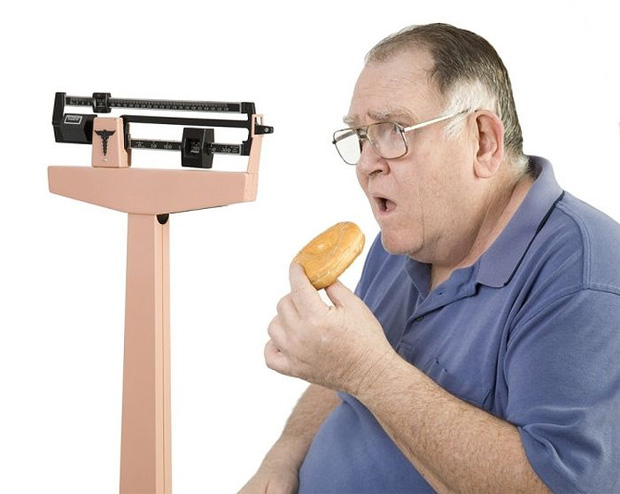 Полный мужчина в очках сидит на фоне медицинских весов и кушает пончик
