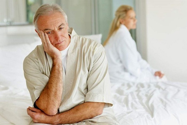 Пожилой мужчина расстроенно сидит на кровати на фоне женщины