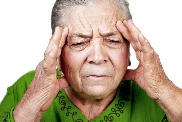Пожилая женщина в зеленой кофте держится двумя руками за больную голову